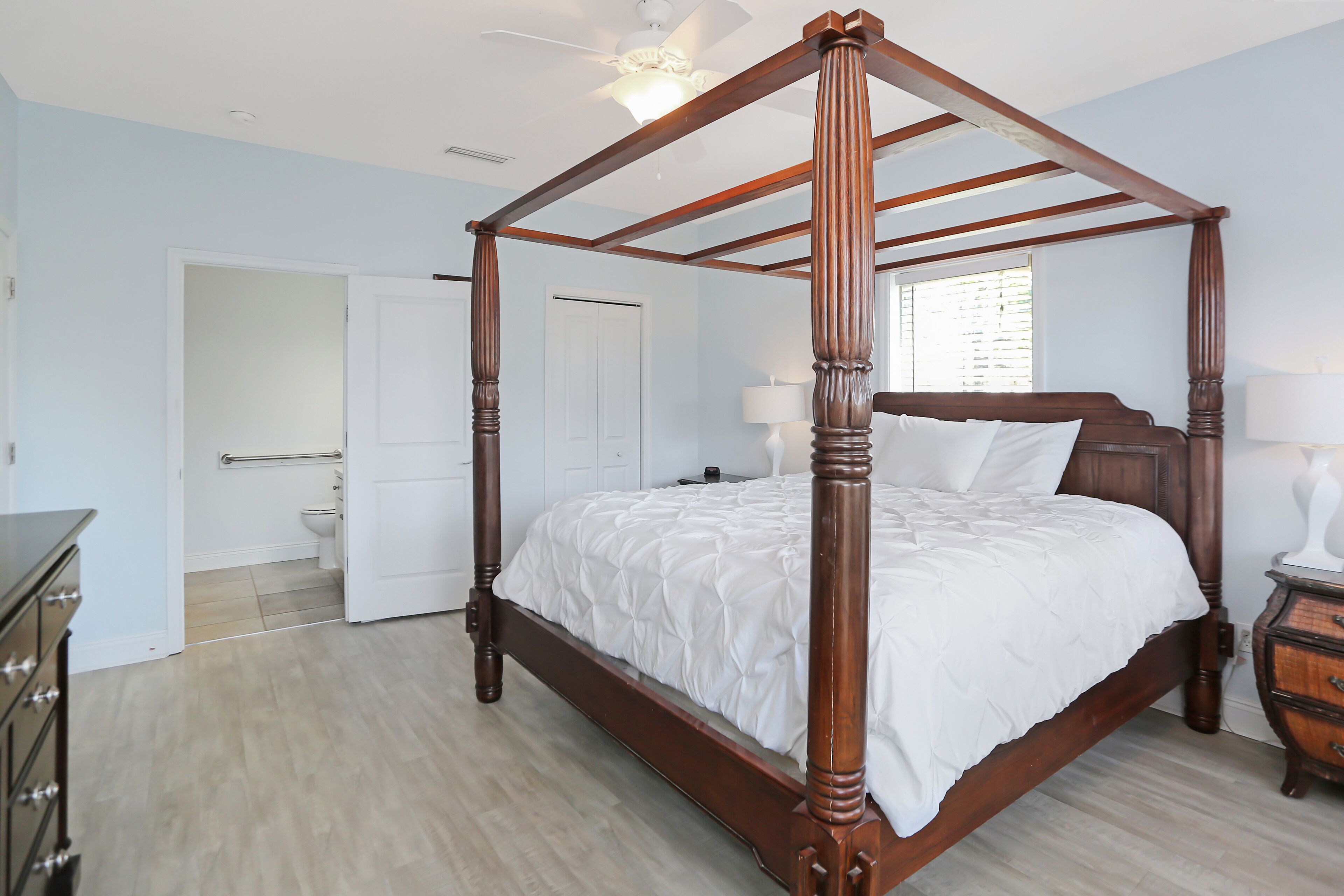 Ein Bett mit Baldachin, dessen Gestell aus Holz besteht, mit weißer Bettwäsche, ein Holzschrank und ein Nachttisch mit Lampe.