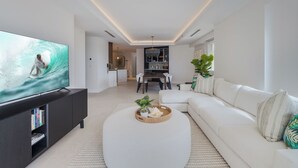 Ein modernes Wohnzimmer mit einem Flachbildfernseher, einem weißen Sofa, einem runden Couchtisch und einem Essbereich im Hintergrund.