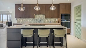Eine moderne Küche mit einer großen Kochinsel, Barhocker, Pendelleuchten und Blick ins Wohnzimmer.