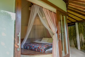 A room with a bed under a canopy, a wooden door, and a view of bamboo outside.
