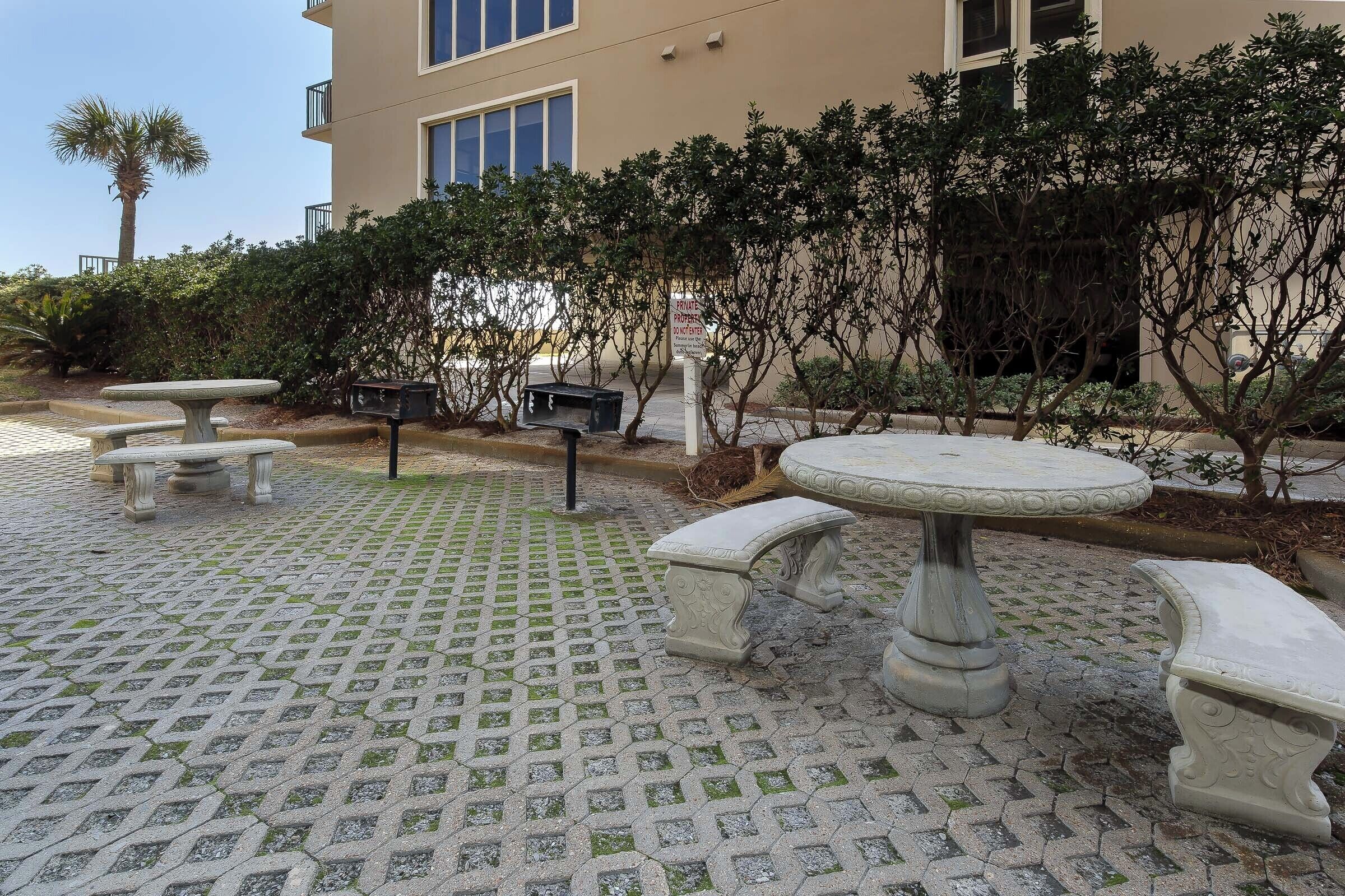 A courtyard with stone tables and benches, surrounded by lush greenery and a building with large windows.