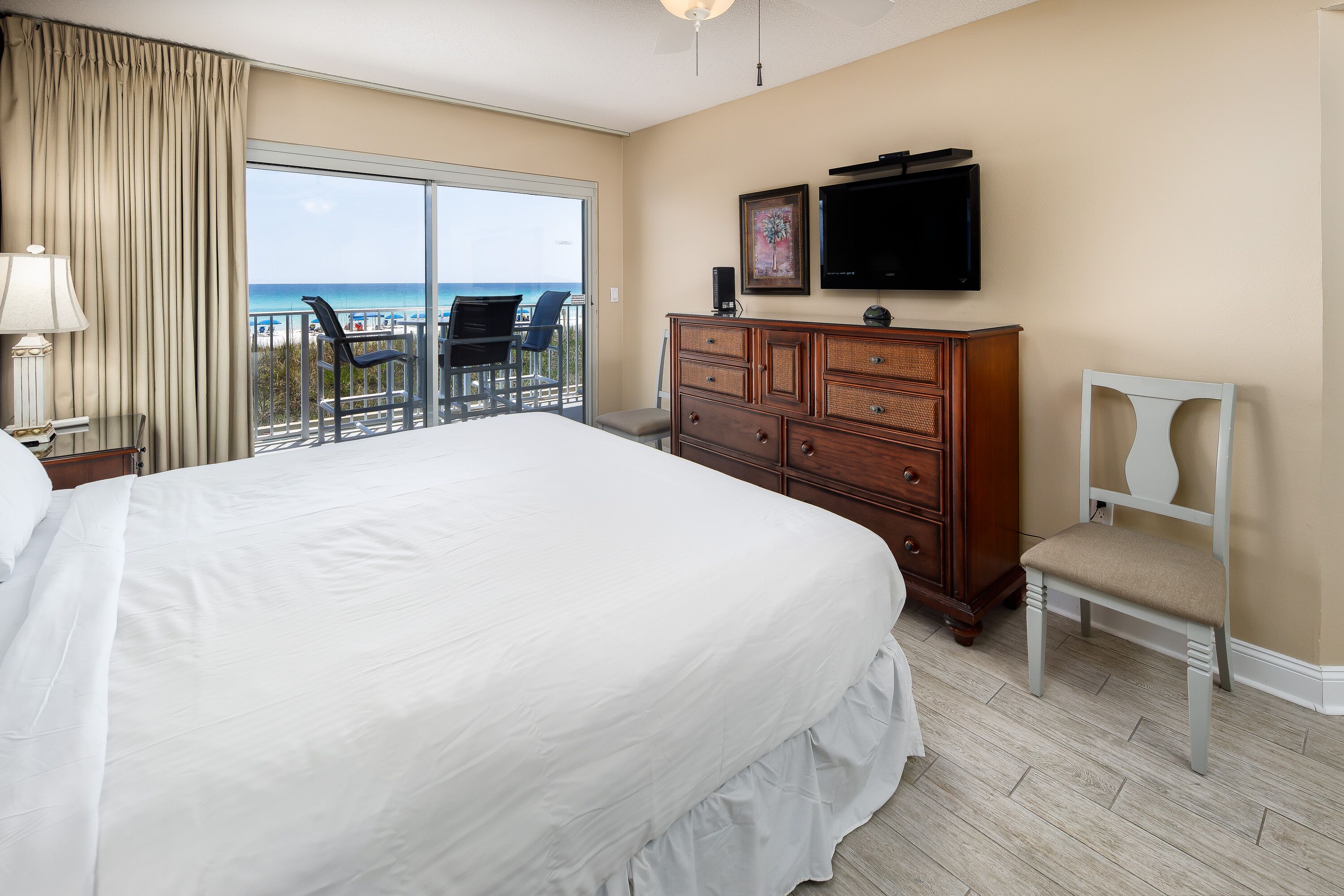 Ein Hotelzimmer mit einem großen Bett, einem Schminktisch, einem Fernseher und einem Balkon mit Blick auf den Strand.
