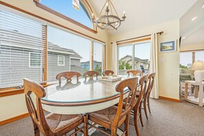 A dining area with a round glass table, wooden chairs, a chandelier, and large windows with blinds.