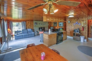 A cozy cabin interior with a wooden ceiling, a dining area, a kitchen with a stove, and a living room with a sofa.