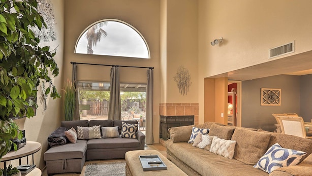 A spacious living room with a large sectional sofa, a small couch, and a coffee table. There is a fireplace, a large window with curtains, and a plant on the left side. - Dog-Friendly Tucson Home w/ Patio & Hot Tub (Tucson)