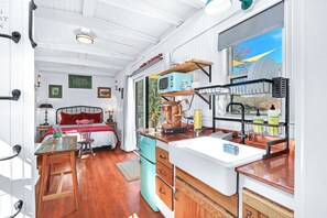 Cocina compacta con pileta, estufa y microondas. Se ve una cama con ropa de cama roja al fondo.
