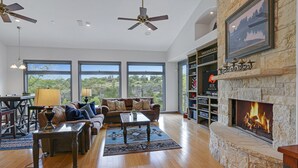 A spacious living area with a stone fireplace, large windows, and a cozy seating arrangement. - Rio Cantando I Blanco River, Pool, Hot Tub, Game Room (Wimberley)