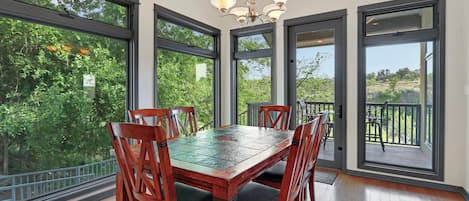 A dining area with a wooden table and chairs, large windows, and a view of greenery.