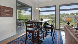 A dining area with a large table and chairs, large windows with a view of greenery, and a wooden sign on the wall. - Rio Cantando I Blanco River, Pool, Hot Tub, Game Room (Wimberley)
