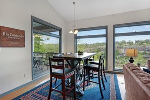 A dining area with a wooden table and chairs, large windows with a view of greenery, and a sign on the wall. - Rio Cantando I Blanco River, Pool, Hot Tub, Game Room (Wimberley)