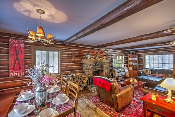 A cozy log cabin interior with a fireplace, wooden furniture, and a dining area with plates and bowls.