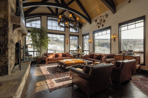 A spacious living room with a fireplace, leather furniture, large windows, and a view of the outdoors. - Stunning Mountain Home by InvitedHome | Hot Tub, Game Room, Golf Course, Hiking (Mountain Village)
