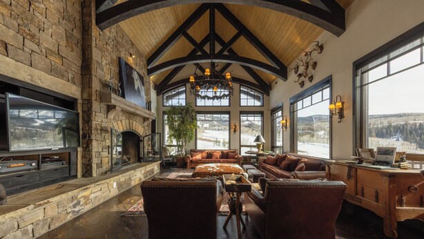 A spacious living area with a stone fireplace, leather furniture, and large windows offering a view of the outdoors. - Stunning Mountain Home by InvitedHome | Hot Tub, Game Room, Golf Course, Hiking (Mountain Village)