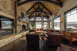 A spacious living area with a stone fireplace, leather furniture, and large windows offering a view of the outdoors. - Stunning Mountain Home by InvitedHome | Hot Tub, Game Room, Golf Course, Hiking (Mountain Village)