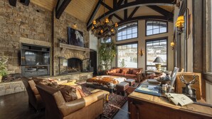 A spacious living room with a stone fireplace, leather sofas, and large windows offering a view of the outdoors. - Stunning Mountain Home by InvitedHome | Hot Tub, Game Room, Golf Course, Hiking (Mountain Village)