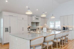 Une cuisine moderne avec des meubles blancs, un grand îlot central doté de tabourets, un éclairage suspendu et une vue sur l’extérieur grâce à de grandes fenêtres.