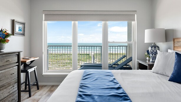 A bedroom with a large bed, a desk, a chair, and a view of the ocean through a balcony.
