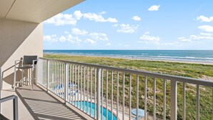 A balcony with a pool, a chair, and a view of the beach and ocean.