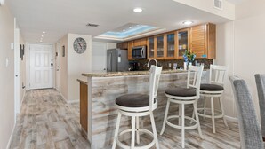 A modern kitchen with wooden cabinets, a central island with bar stools, a clock on the wall, and a ceiling light.