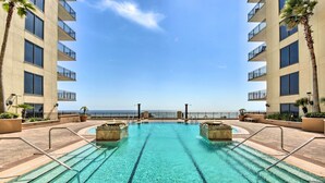 Piscina com água azul cristalina, rodeada por um deck de tijolos e com vista para uma praia.