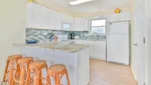 A modern kitchen with white cabinets, a central island with stools, a refrigerator, and a window providing natural light. - Krabby Patty – Oceanfront Family Fun (Topsail Beach)