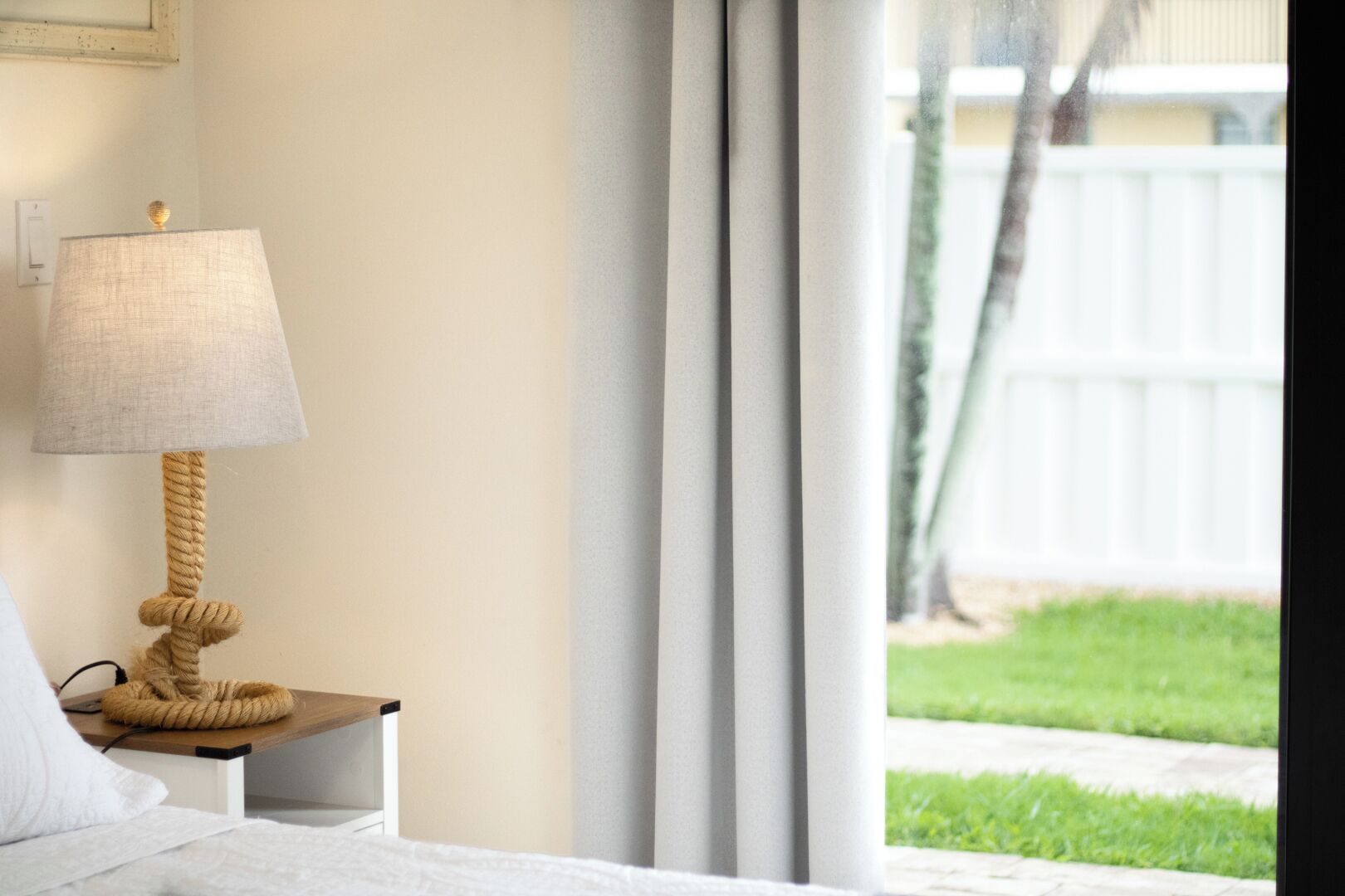 A bedroom with a bed, a bedside table with a lamp, and a view of a white fence and greenery outside.