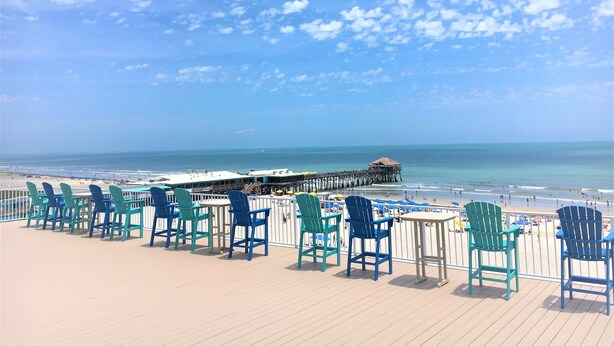 Um terraço com cadeiras e mesas com vista para uma praia e um cais.