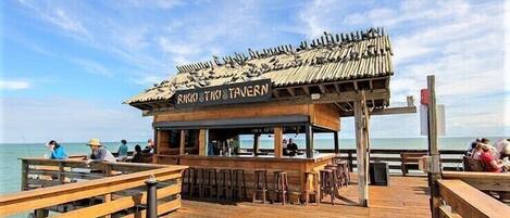 Een houten steiger die uitkomt bij een rustieke taverne met rieten dak en een bar.