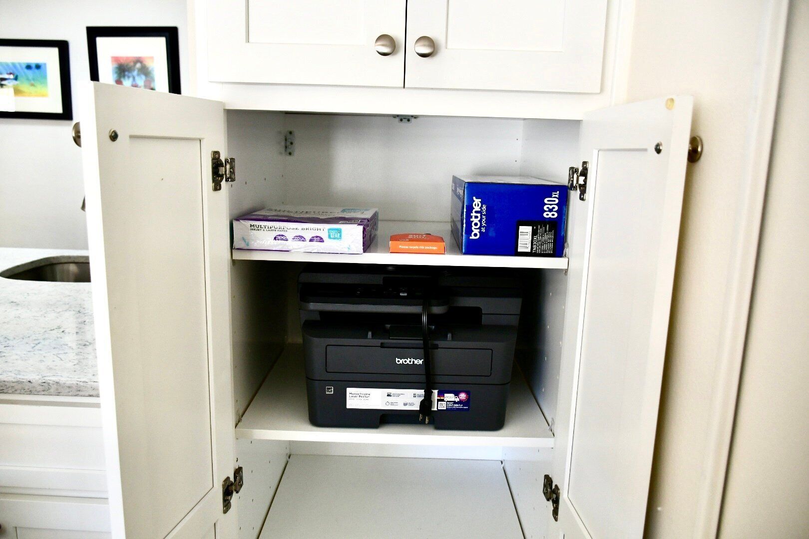 A storage cabinet with a printer, paper, and a box labeled "Brother" in a kitchen setting.