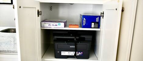 A storage cabinet with a printer, paper, and a box labeled "Brother" in a kitchen setting.