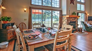 A dining area with a wooden table set for four, surrounded by white chairs with cushioned seats. A stone fireplace with a wooden mantel is visible in the background.