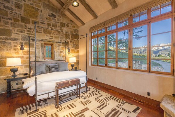 A bedroom with a stone wall, a large window with a mountain view, a four-poster bed, and a patterned rug. - Basque-Influence Home by InvitedHome | Gourmet Kitchen, Winter Paradise, Hot Tub (Telluride)