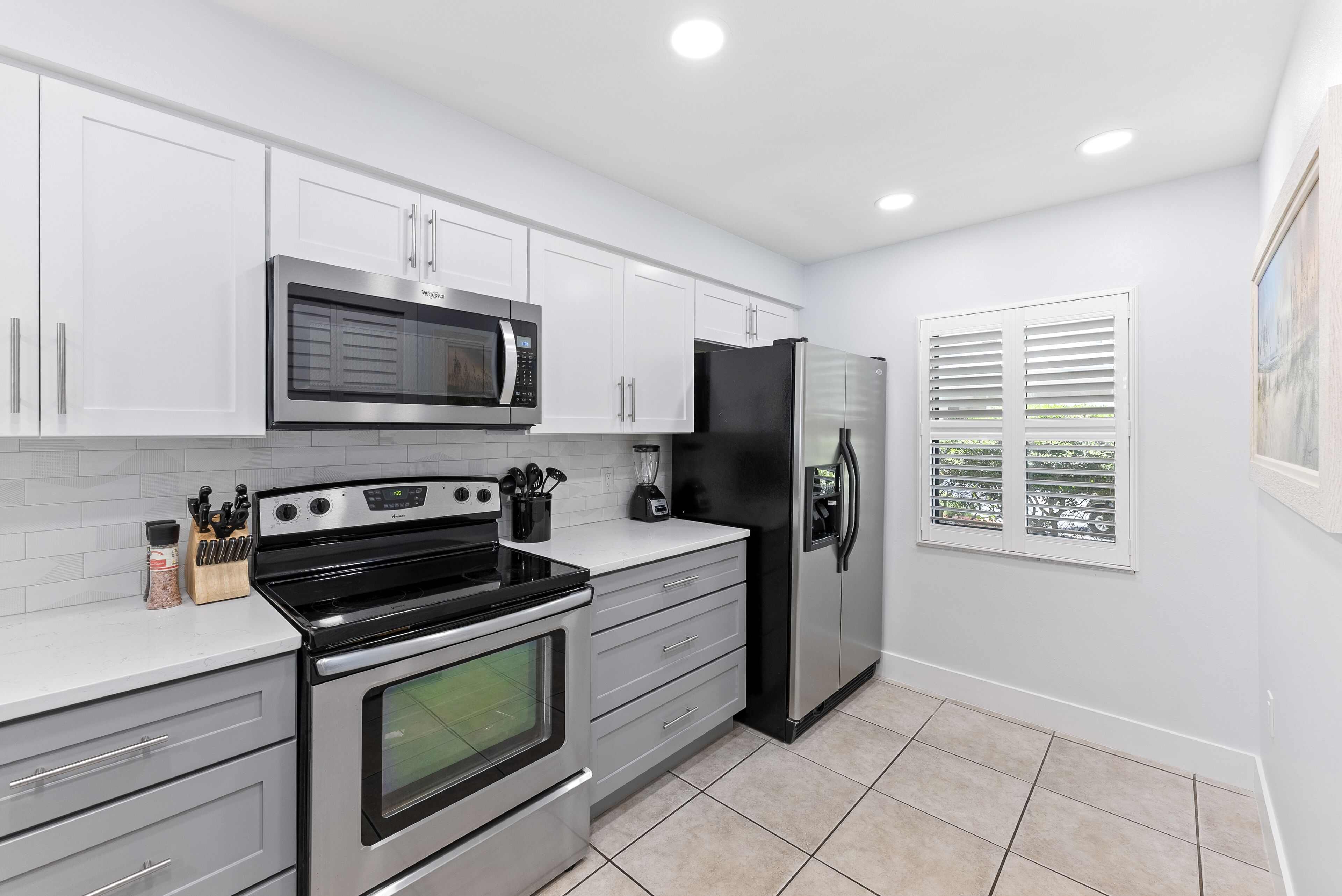 A modern kitchen with white cabinets, stainless steel appliances, and a window with blinds.