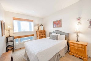 A bedroom with a large bed, bedside tables, a dresser, a window with blinds, and wall art.