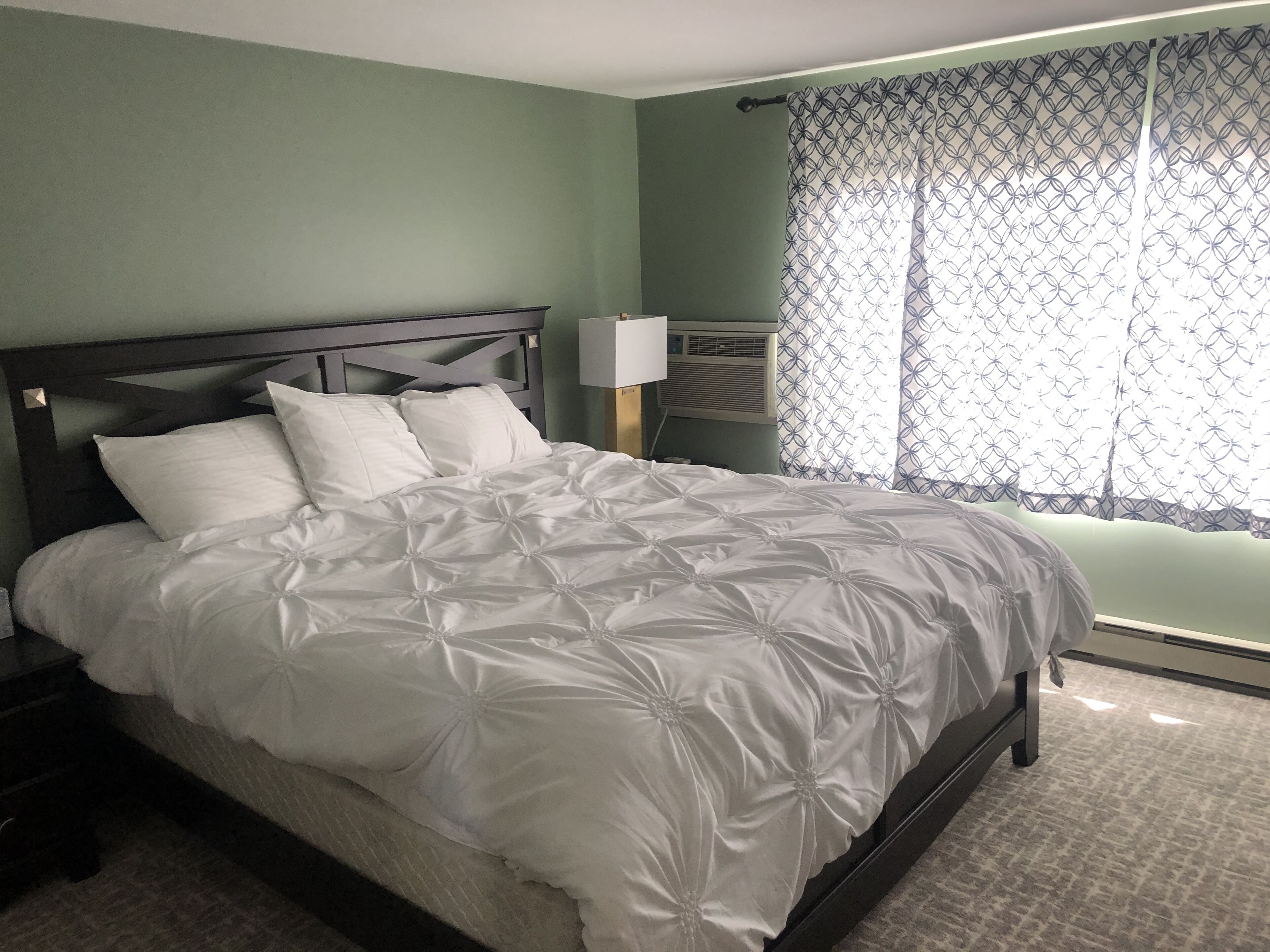 A neatly made bed with a white duvet and pillows, a dark wooden headboard, a bedside table with a lamp, and a window with patterned curtains.