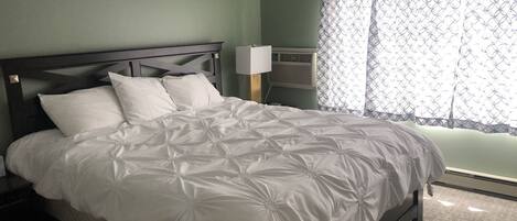 A neatly made bed with a white duvet and pillows, a dark wooden headboard, a bedside table with a lamp, and a window with patterned curtains.