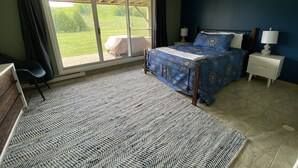 A bedroom with a large sliding glass door leading to a patio, a bed with a blue patterned bedspread, a nightstand with a lamp, and a patterned rug. - Leelanau County, 4-brm 4-bath. A/C, Pool, 5 mins to Lk Michigan Beach (Cedar)