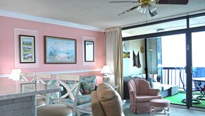 A room with a pink wall, a dining area with a table and chairs, a sofa, and a view of the outdoors through a glass door. - Comfortable 2 bedroom oceanfront condo popping with color that has free WiFi, and a balcony with an amazing ocean view located uptown just steps to the beach! (Ocean City)