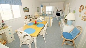 Una habitación acogedora y bien iluminada con una mesa de comedor de madera, sillas blancas y una silla de mimbre. Se ve un pequeño espacio de cocina al fondo.
