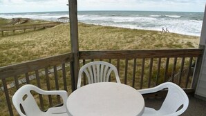Un área de estar al aire libre techada, con una mesa y sillas, con vista a una playa con olas y una duna cubierta de pasto.