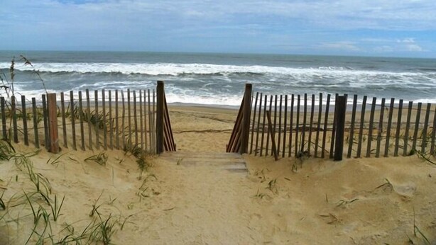 Una entrada a la playa con una valla de madera y un camino arenoso que lleva al océano.