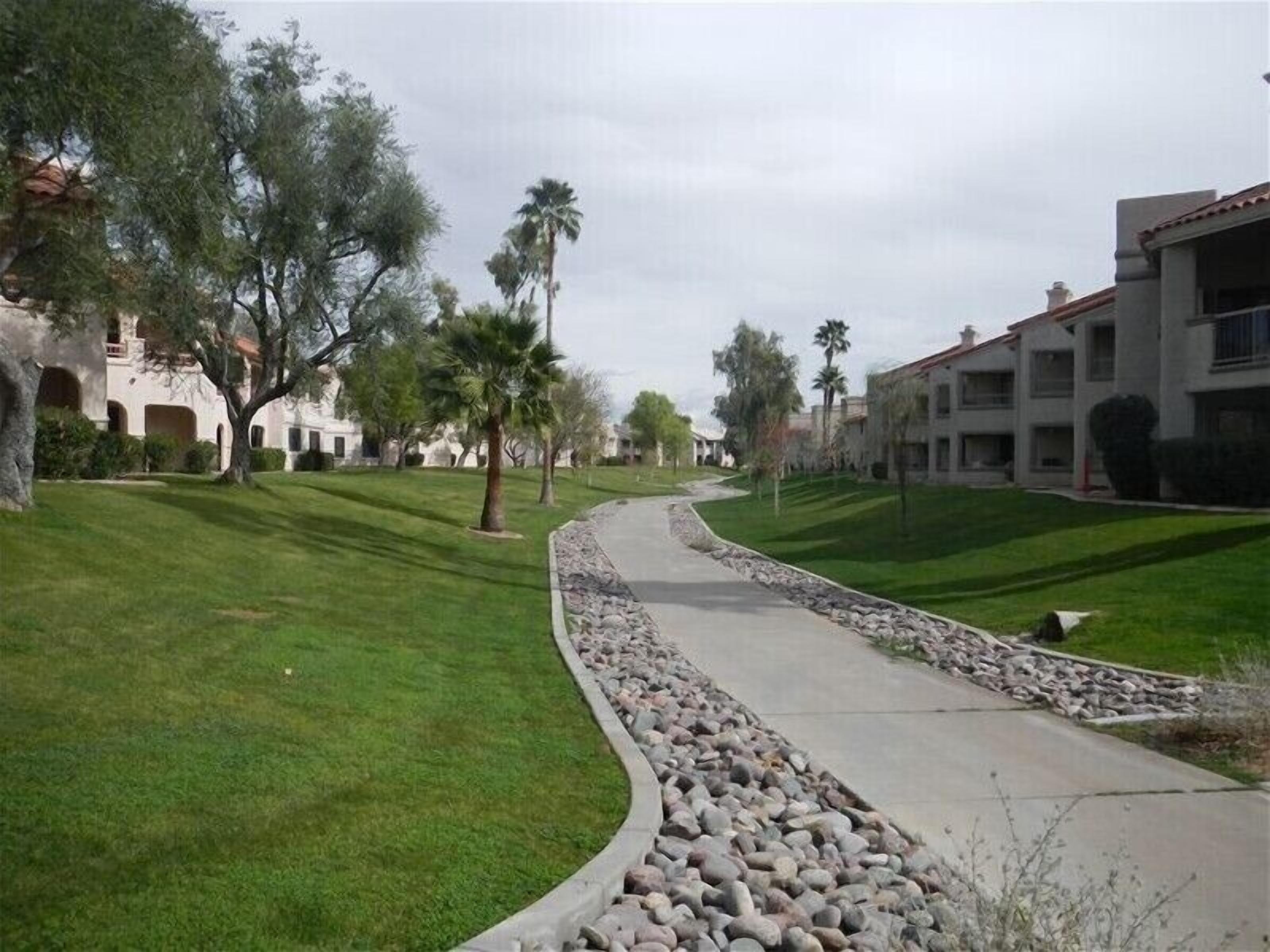 A pathway curving through a well-maintained lawn with a stone border, flanked by trees and buildings with a Mediterranean architectural style.