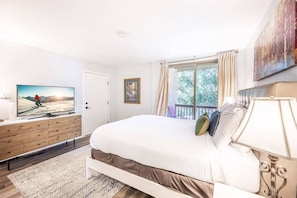 Une chambre à coucher avec un lit, un téléviseur posé sur une commode, une fenêtre avec des rideaux et un tableau accroché au mur.