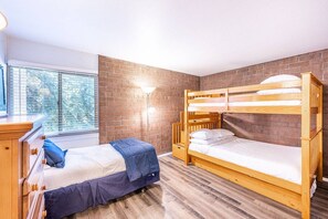 Une chambre avec un lit superposé, un lit simple, un bureau avec des tiroirs, une lampe et une fenêtre avec des stores.