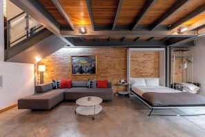 A modern hotel room with a lofted sleeping area, a sofa, a small table, and a brick wall with a painting.