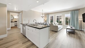 A spacious kitchen with white cabinetry and a black countertop, a dining area with a table and chairs, and a living room with a sofa and a TV. - Elegant 2BR | Pool, Lake Views | Remodeled June 24 (Henderson)