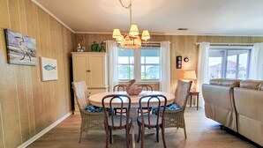 A dining area with a round table, four chairs, a chandelier, and a view of the outside through a window. - Merri Mac – Breezy Stays by the Sea (Surf City)