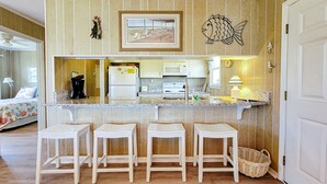 A kitchen with a wooden panel backsplash, a framed picture, and a fish decoration. There are three white stools in front of the counter. - Merri Mac – Breezy Stays by the Sea (Surf City)