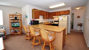A compact kitchen with wooden cabinets, a built-in microwave, a refrigerator, and a dining area with wicker chairs. - Spacious, quaint 2 bedroom condo with free WiFi and a large balcony located downtown on the bayside just two blocks from the beach! (Ocean City)