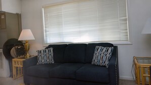 A dark blue sofa with patterned pillows, a small table with a lamp, and a window with blinds. - Spacious, quaint 2 bedroom condo with free WiFi and a large balcony located downtown on the bayside just two blocks from the beach! (Ocean City)